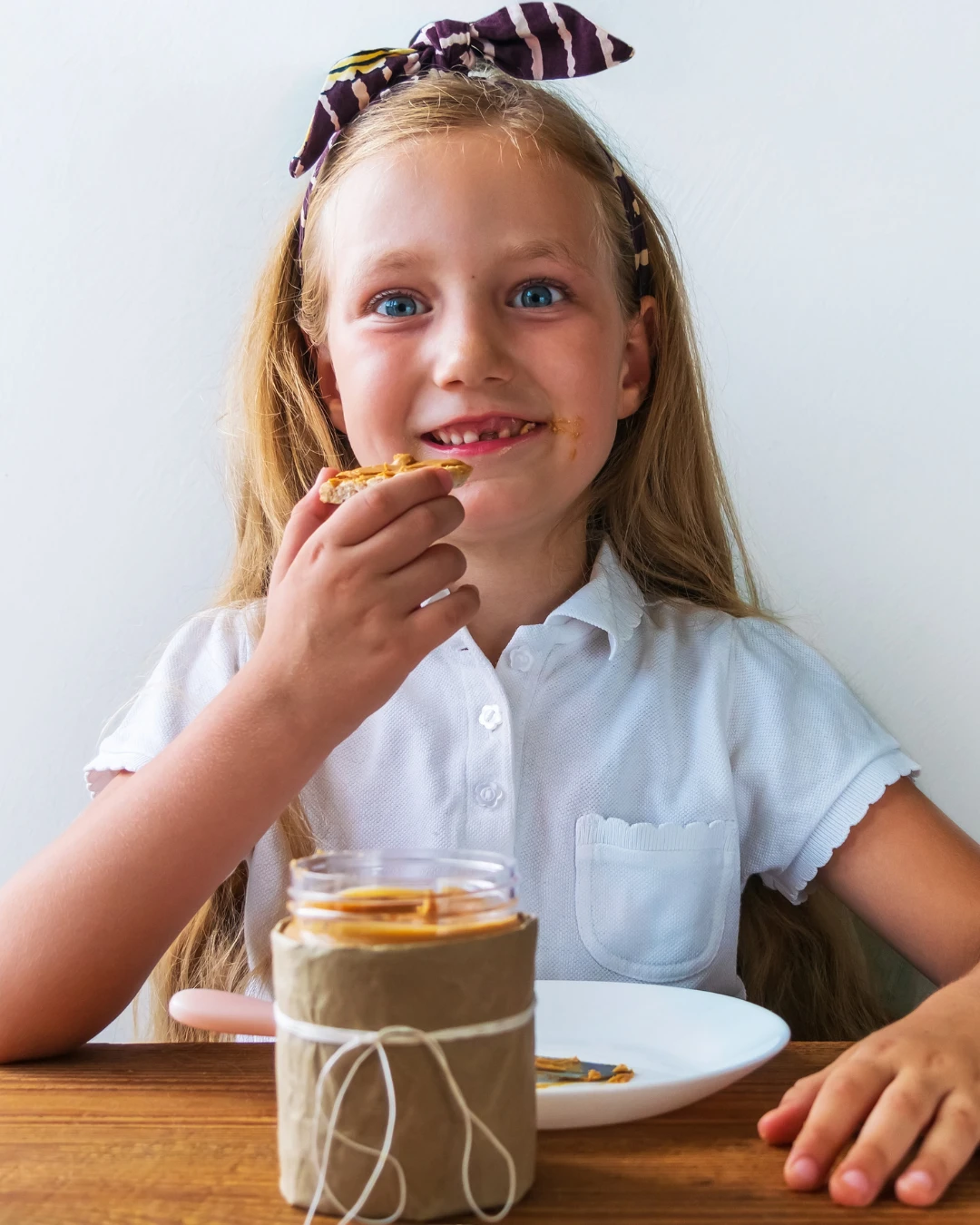 A smiling girl eating a cracker with peanut butter at a table, the jar in front of her